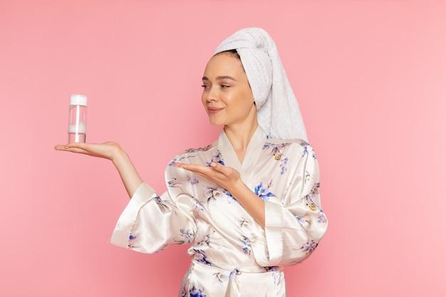 Woman in bathrobe with towel on head presenting skincare product on pink background