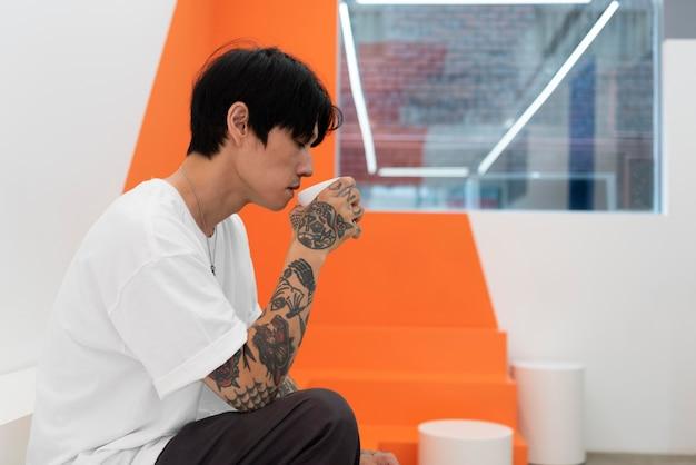 Man with tattoos sitting thoughtfully in a modern gallery space with orange sculptures